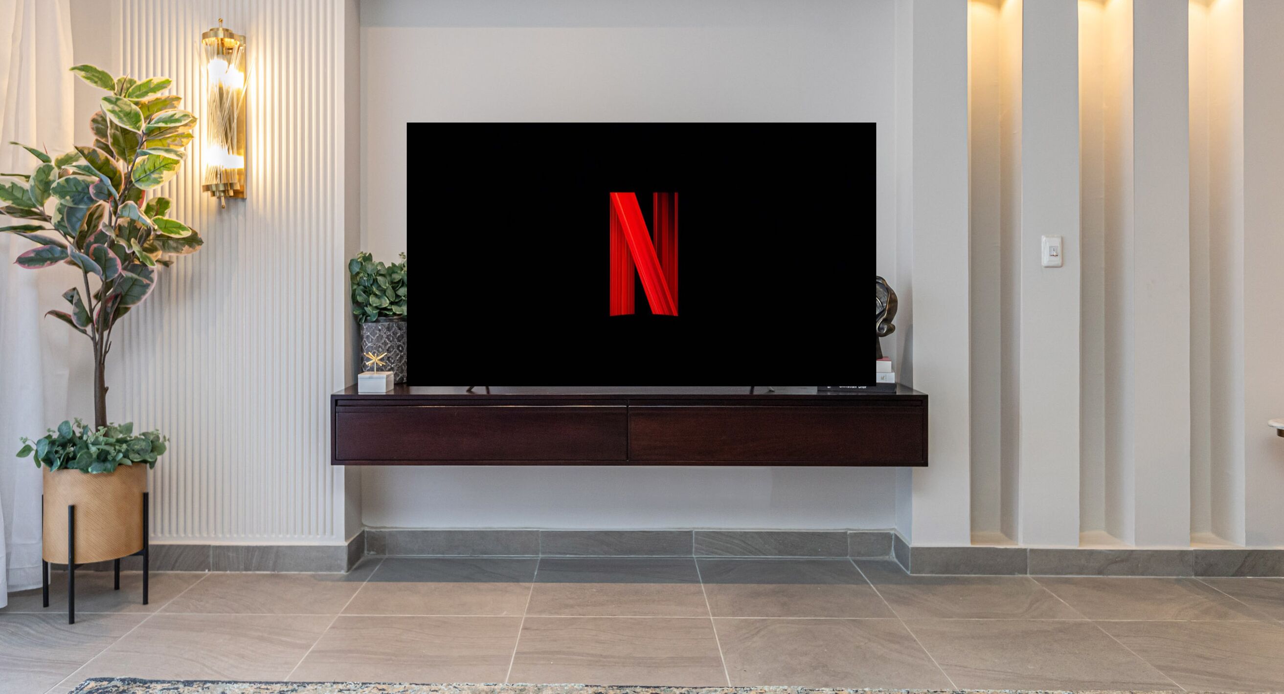 A modern living room with a flat-screen TV mounted on a dark wooden console, a potted plant, and a decorative wall light.