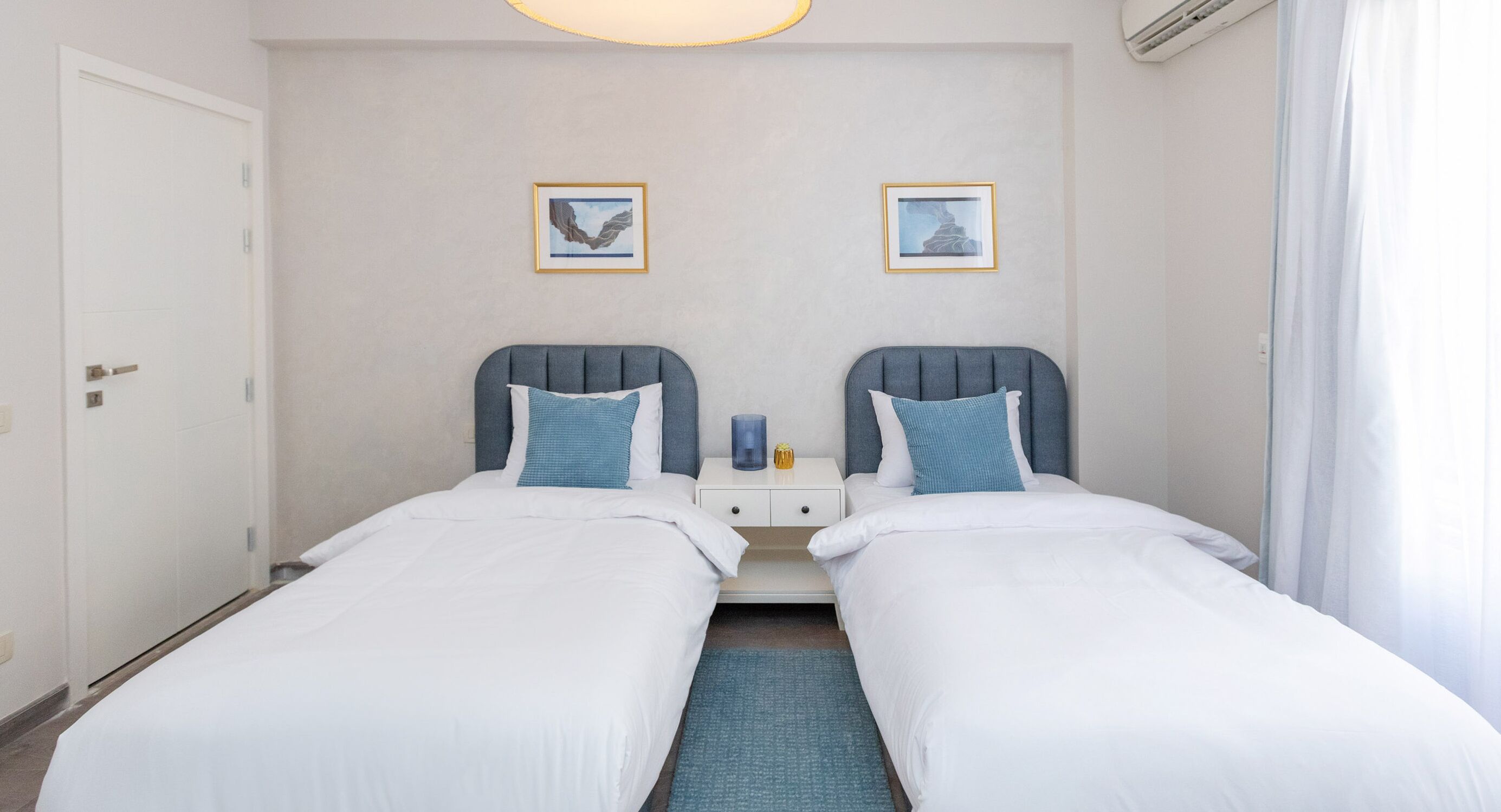 A hotel room with two single beds, a nightstand with a lamp, and two framed pictures on the wall.
