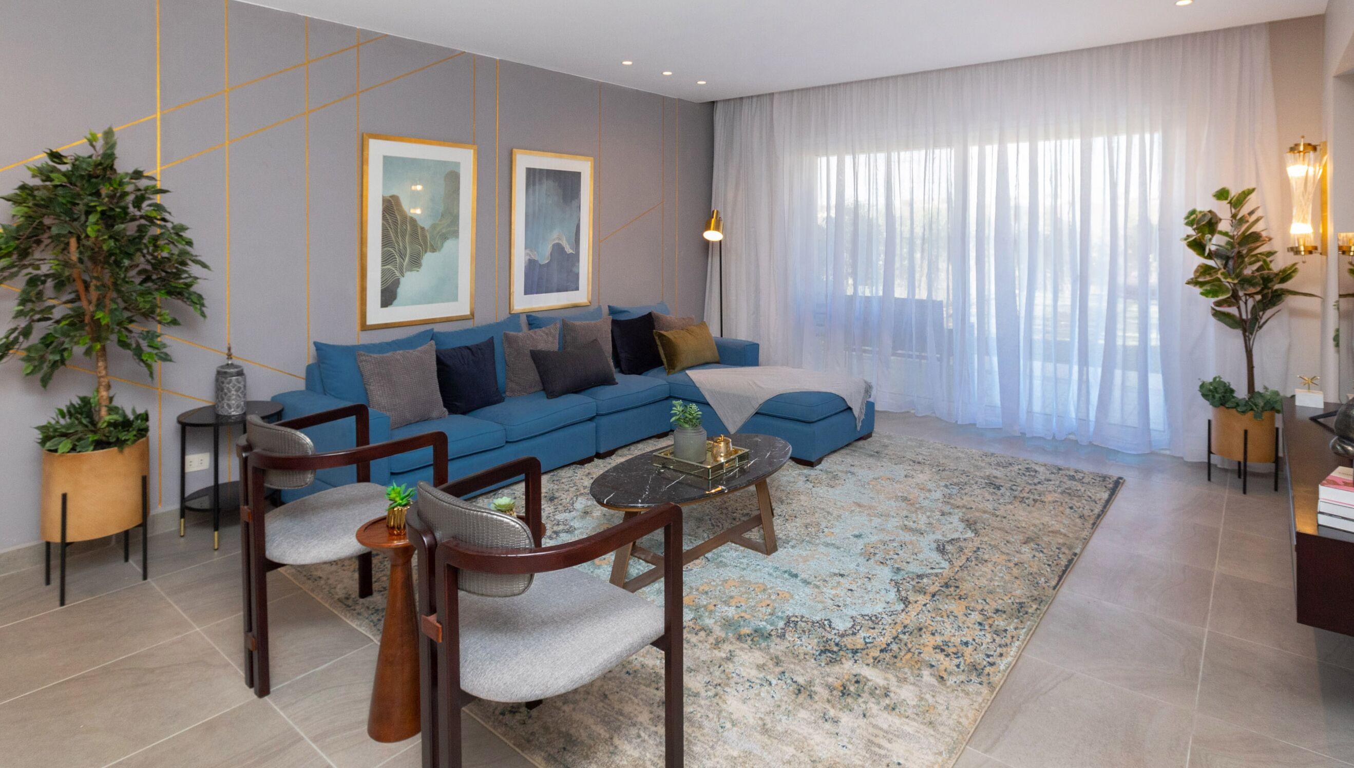 A modern living room with a blue sofa, two framed artworks, a coffee table, and a large window with sheer curtains.