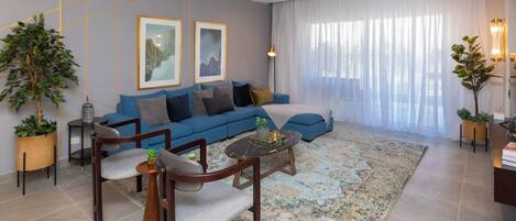 A modern living room with a blue sofa, two framed artworks, a coffee table, and a large window with sheer curtains.