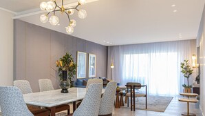 Un área de comedor moderna con una mesa de mármol, sillas estampadas, una lámpara de araña y vistas del salón a través de un ventanal grande.