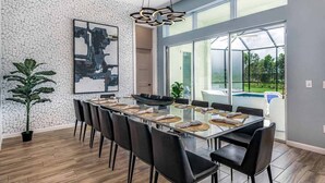 A modern dining area with a long table, black chairs, a large abstract painting, and a view of a swimming pool through large windows. - Trendy Home w Trampoline RM, Bowling, Fantasy Themed BR & Theater - Near Disney (Davenport)