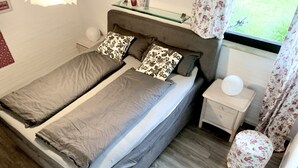 En dubbelsäng med grå sängkläder, ett nattduksbord med en vit lampa och ett fönster med blommiga gardiner.