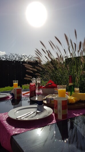 An outdoor dining table with plates, cutlery, and drinks, set against a backdrop of tall grasses and a clear sky.