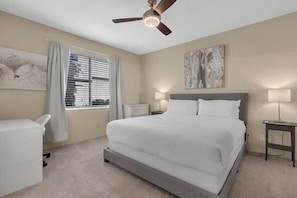 A bedroom with a bed, ceiling fan, bedside lamps, a nightstand, a window with blinds, and a wall-mounted artwork.