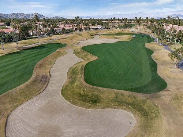 A golf course with sand bunkers, a putting green, and a view of a residential area in the background.