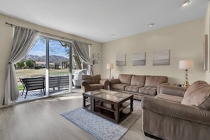 A spacious living room with a large sliding glass door leading to a patio, a leather sofa set, a glass coffee table, and wall-mounted lamps.