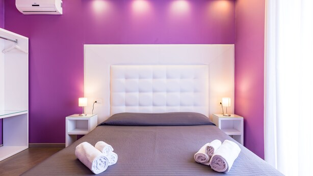 Een moderne hotelkamer met een paarse wand, een groot bed met een gestuikt hoofdeinde, twee nachtkastjes met lampen, en opgerolde handdoeken op het bed.