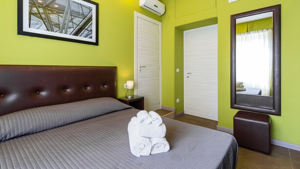A hotel room with a bed, a stack of towels, a framed picture, a mirror, and a wardrobe. - BH Napoli Railway - Pistacchio - 4 (Napoli)