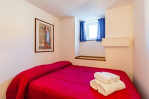 A bedroom with a red bedspread, a framed artwork on the wall, a window with blue curtains, and a white towel on the bed. - Parthenope's Eye on the Seafront (Napoli)
