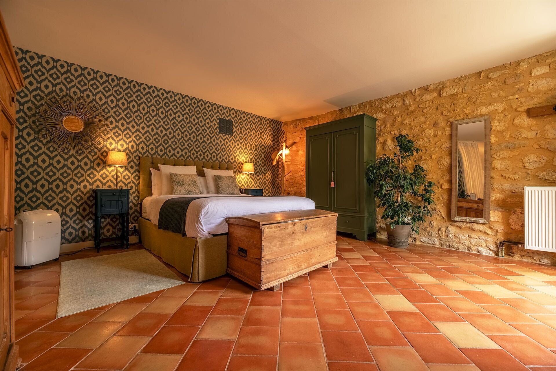 Un dormitorio con pared estampada, una cama con un cofre de madera, una puerta verde, una planta en maceta y un radiador.