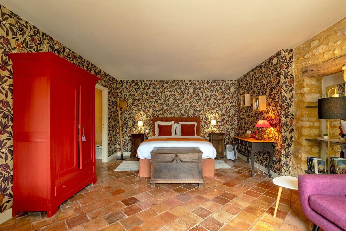 Une chambre avec une grande armoire rouge, un lit avec une literie orange, une commode en bois, une petite table avec une lampe et un mur en pierre.