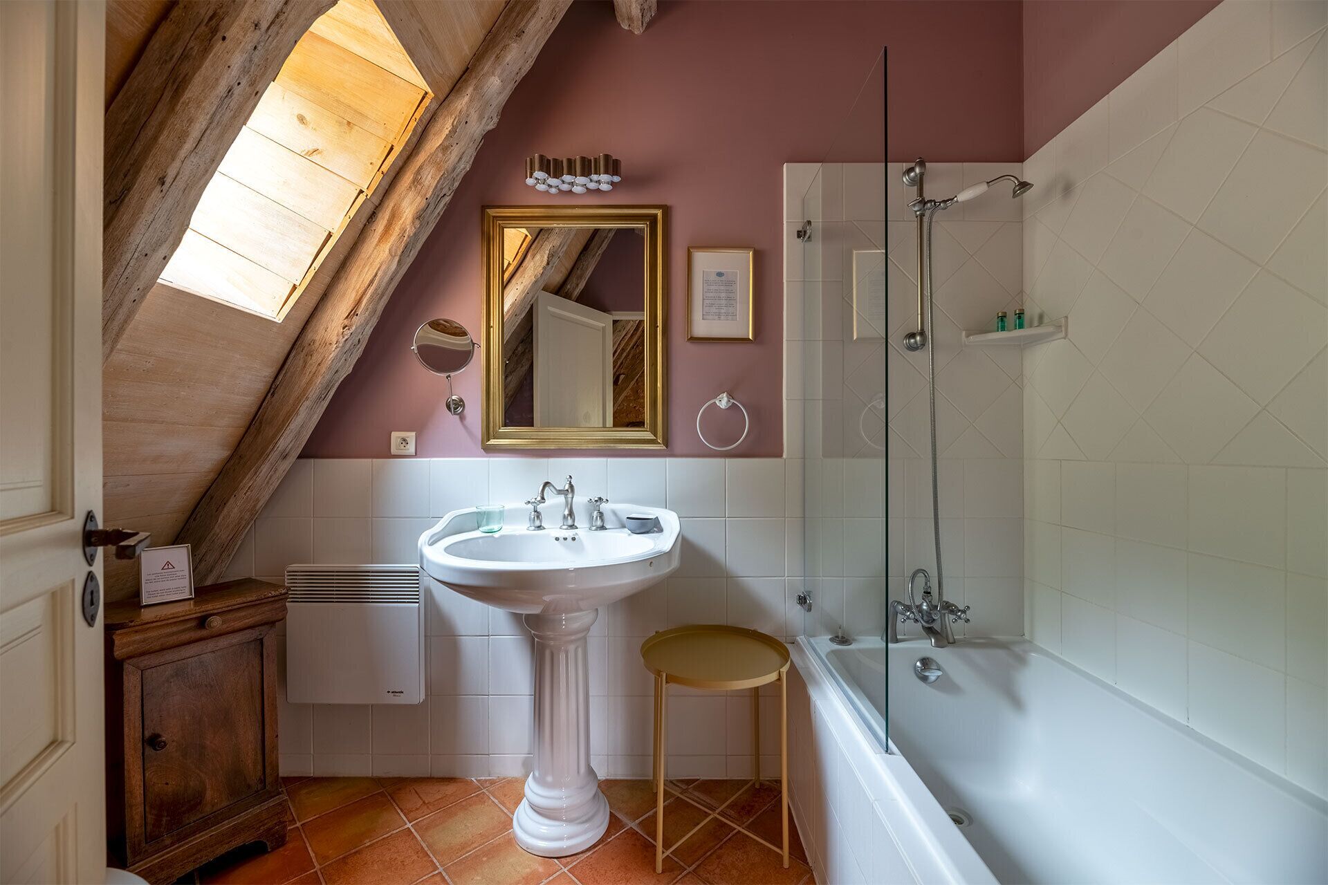 Een badkamer met een wastafel op voet, een spiegel, een douche en een houten kruk.