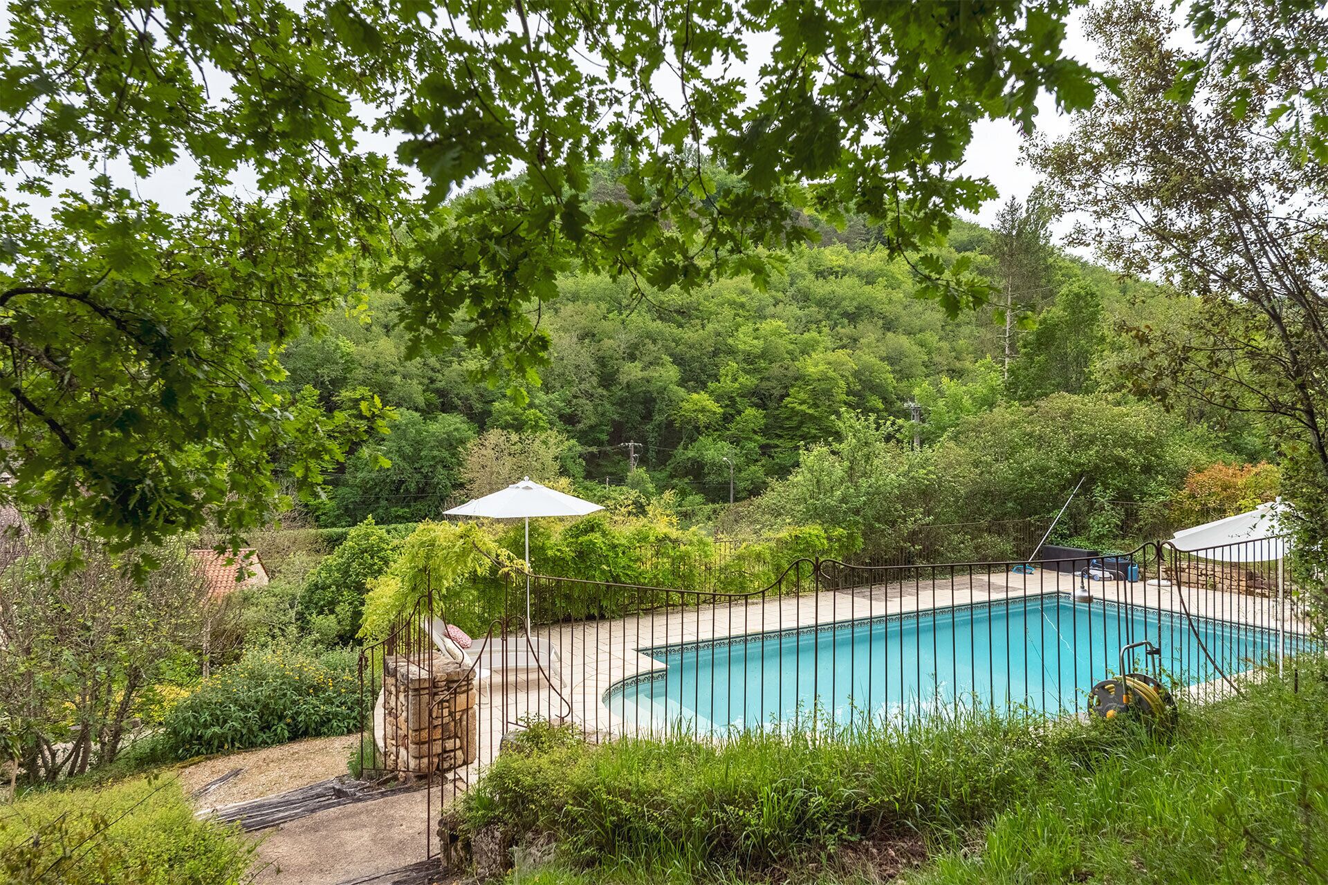 A rectangular swimming pool surrounded by a metal fence, with a stone structure and a small garden area.
