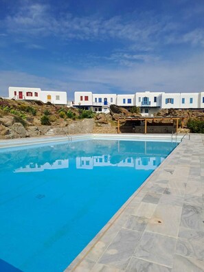 A swimming pool with a clear blue water surface, surrounded by a stone deck. In the background, there are white buildings with blue accents and red doors. - Amazing view of Ornos Bay in Mykonos (C869+V7P Mikonos, Greece)