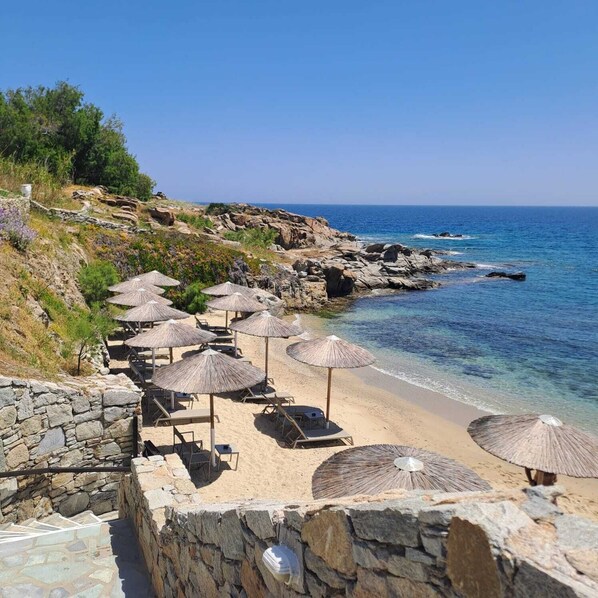 A beach with stone walls, umbrellas, and lounge chairs. - Amazing view of Ornos Bay in Mykonos (C869+V7P Mikonos, Greece)