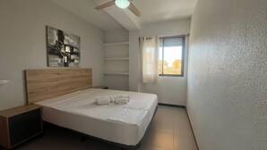 A bedroom with a wooden headboard, a bed with white linens, a nightstand, a ceiling fan, a window with curtains, and a wall-mounted abstract art piece. - Tamaris playa 7ºC Solo Familias! (Xeraco Playa)