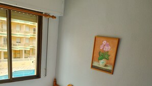 Una habitación con dos camas individuales, una mesita de noche de madera, un cuadro colgado en la pared y una ventana con vista a un edificio.