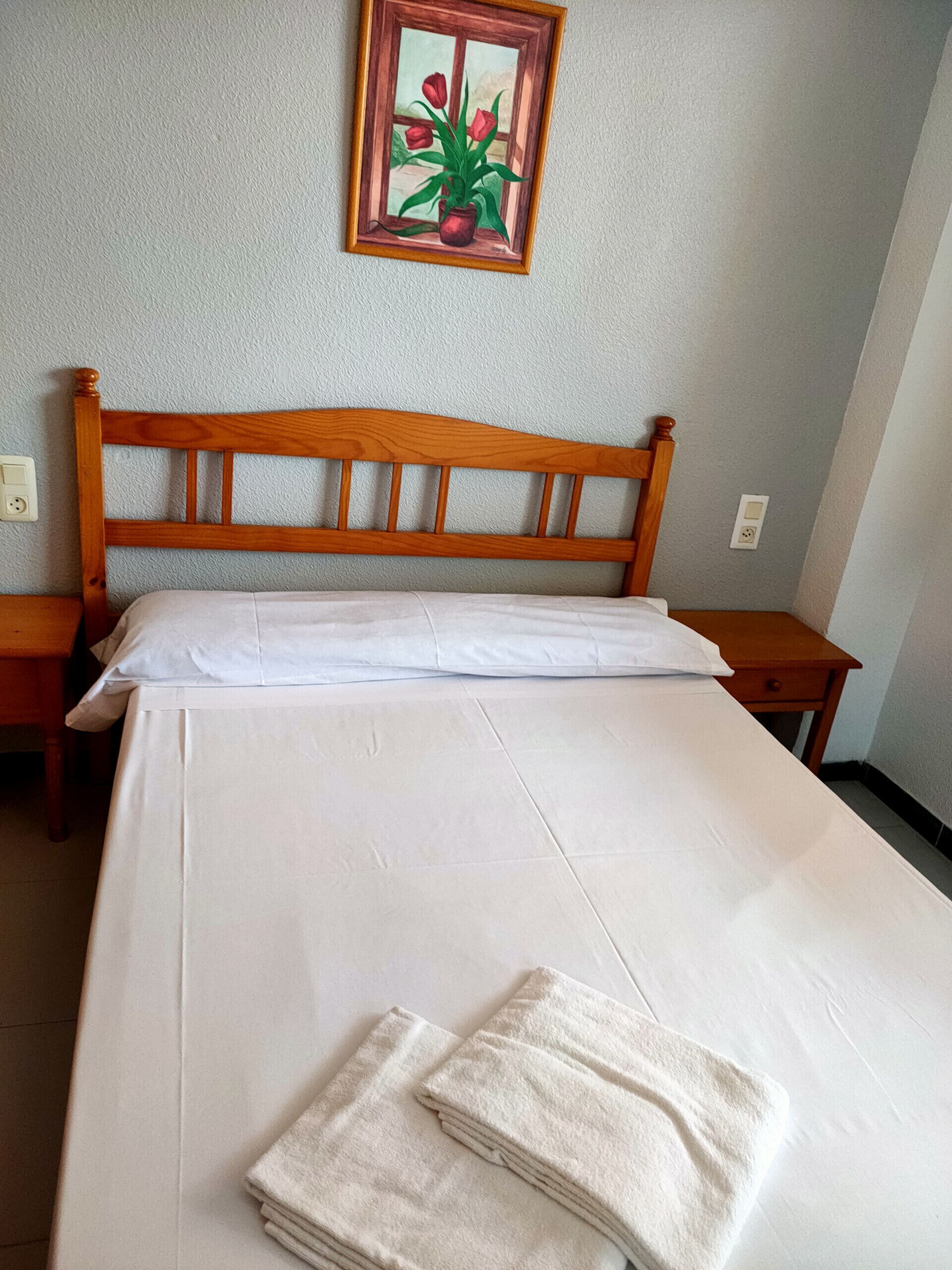 Een net opgemaakt bed met witte lakens en een houten hoofdbord, een klein nachtkastje en een schilderij van bloemen aan de muur.