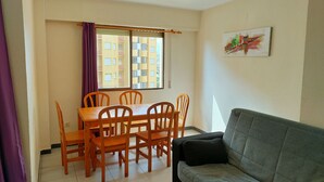 Una sala de estar con un sofá gris, mesa de comedor de madera con seis sillas, una ventana con vistas a edificios y un cuadro en la pared.
