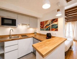 Cozinha moderna com micro-ondas, pia e bancada de madeira. Há armários brancos, uma pequena planta e uma máquina de café. O quarto tem teto de madeira e uma cama com manta branca.