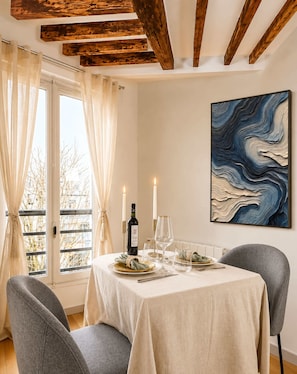 Uma área de refeições com uma mesa posta para dois, uma garrafa de vinho, duas velas e um quadro azul abstrato na parede.