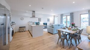 A modern kitchen with a central island, a dining area with a table and chairs, and a living room with a sofa. - 2 Deben Wharf, Woodbridge (Woodbridge)