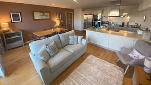 A modern living room with a sofa, a dining table, and a kitchen area with a refrigerator and stove. - 2 Deben Wharf, Woodbridge (Woodbridge)