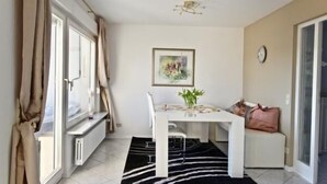 Une petite pièce lumineuse, avec une table blanche, une chaise, un tapis noir et blanc, une fenêtre avec des rideaux, un radiateur, une horloge et un tableau encadré au mur.