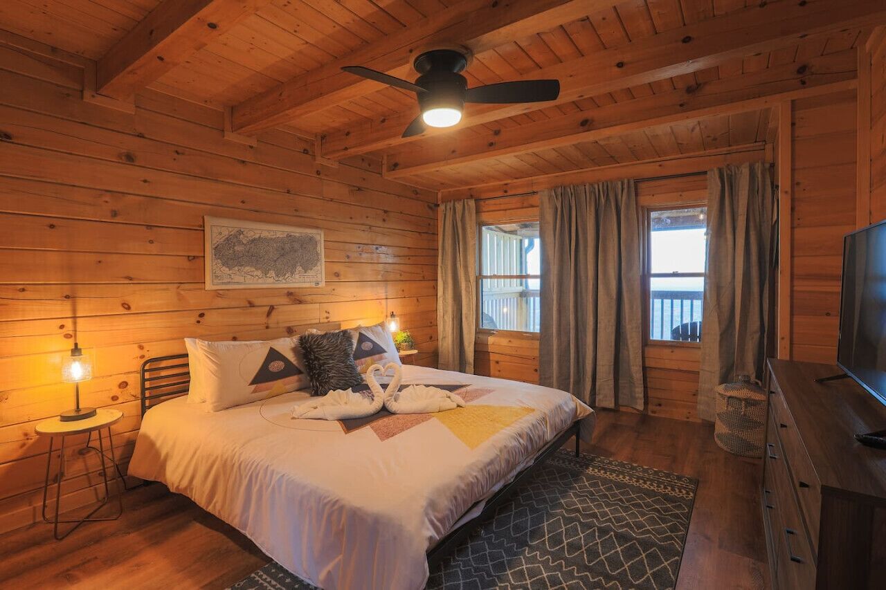 A wooden cabin bedroom with a bed, a ceiling fan, a television, and a window with curtains.