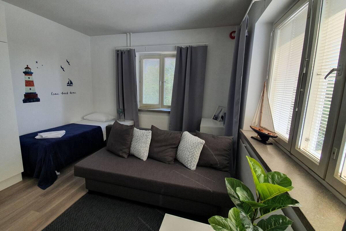 Une pièce de taille modeste comprenant un canapé, un petit lit, une fenêtre avec des stores, une plante et un élément de décoration murale.