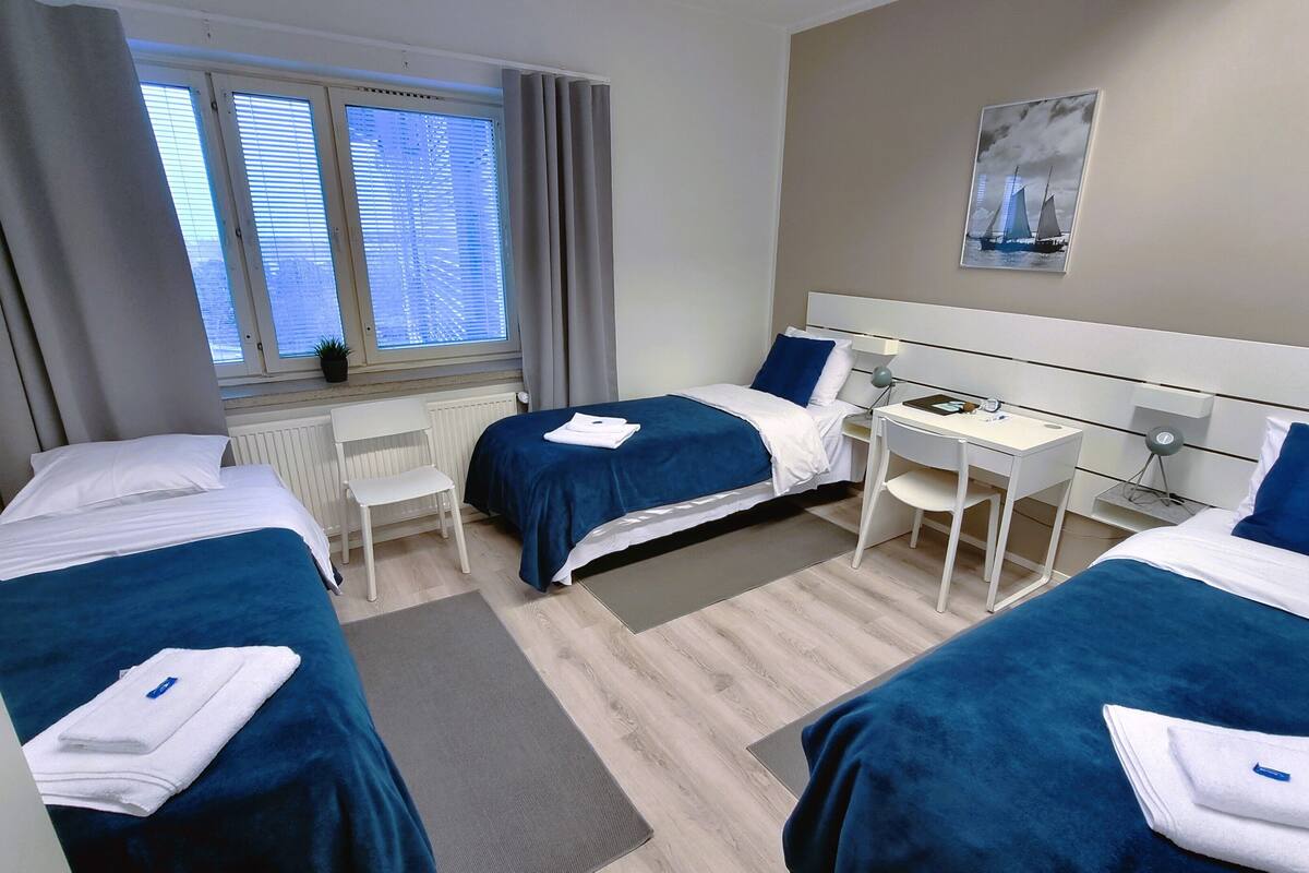 Une chambre d’hôtel avec deux lits, un bureau, une chaise, une fenêtre avec des stores, un radiateur et un tableau au mur.