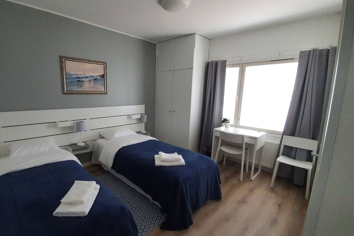 Une chambre d’hôtel avec deux lits, un bureau, une chaise, une fenêtre avec des rideaux et un tableau au mur.