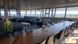 Un vaste espace intérieur doté d’une longue table en bois et de plusieurs canapés en cuir noir, de grandes fenêtres offrant une vue sur l’eau et les collines, et d’un design de plafond moderne.