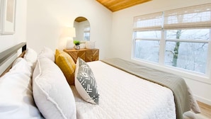 A bedroom with a wooden ceiling, a large window, a bed with white bedding, a bedside table with a lamp, and a mirror. - The Aerie ~ A Modern Retreat at Double Bridges (Mountain View)