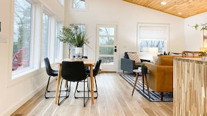 A spacious room with a wooden ceiling, large windows, a dining area with a table and chairs, and a seating area with a sofa and armchairs. - The Aerie ~ A Modern Retreat at Double Bridges (Mountain View)