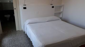 Una cama individual con ropa de cama blanca en una habitación con piso de baldosas y una puerta blanca.
