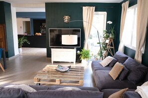 A living room with a dark green wall, a flat-screen TV, a wooden coffee table, a grey sofa, and a potted plant.