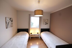 A room with two single beds, a small bedside table with lamps, a window with curtains, and wall art.