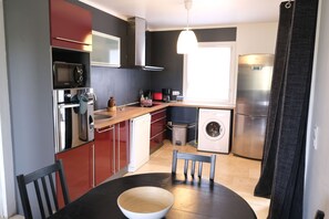 A compact kitchen with a microwave, oven, and dishwasher. There is a dining table with chairs and a bowl on it.