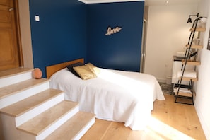 A bedroom with a bed, a wooden headboard, a white bedspread, a blue accent wall, a small orange lamp, and a wooden staircase.