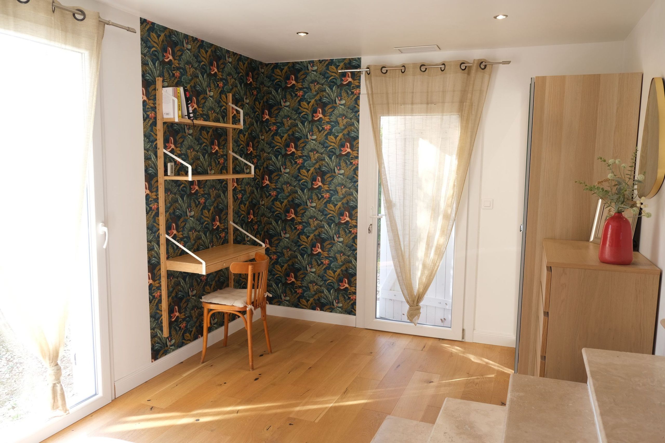 A room with a wooden floor, a bookshelf, a chair, a window with curtains, and a mirror on the wall.