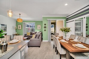 A modern kitchen with a dining area, featuring a wooden dining table set for four, a grey sofa, and a kitchen island with a sink and counter. - Lovely Townhome w/ Private Splash Pool + FREE Resort Access! (Kissimmee)