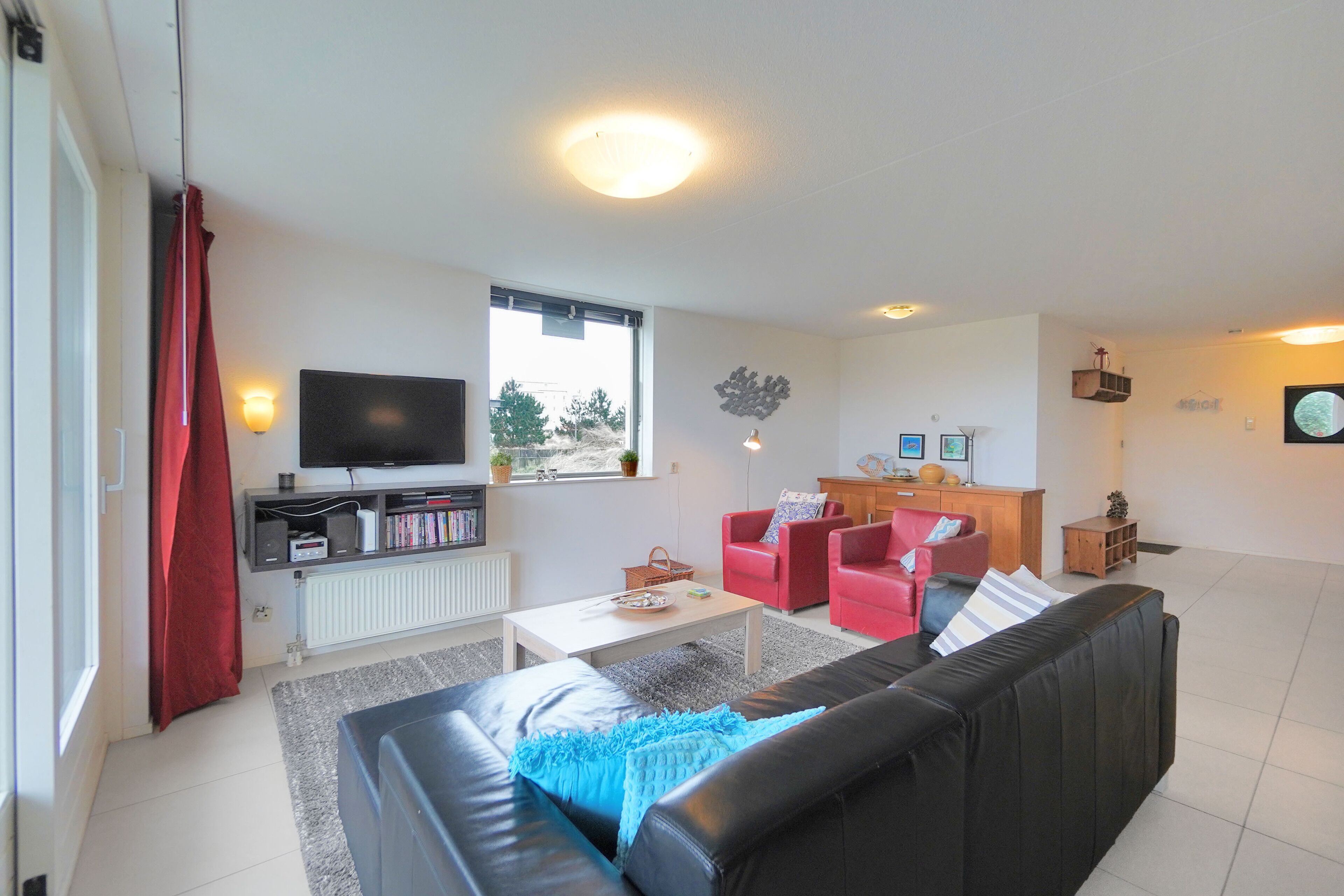 Een woonkamer met een zwarte bank, rode stoelen, een witte salontafel en een televisie op een dressoir.