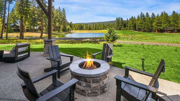 Un área de estar al aire libre con un hogar de piedra rodeado de sillas Adirondack, con vistas a un lago y un paisaje boscoso.
