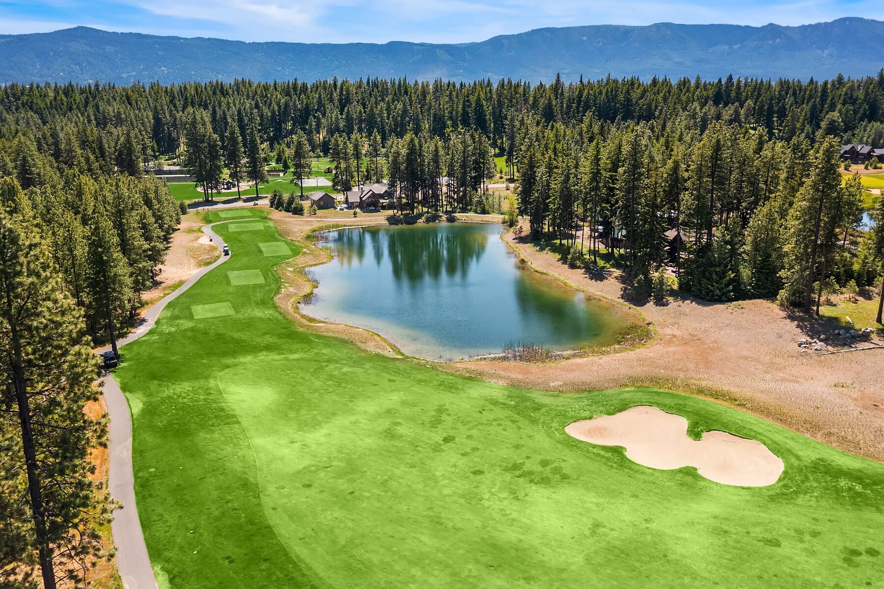 A golf course with a pond, surrounded by dense forest and mountains in the background.