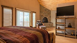 A bedroom with a large bed, a stone fireplace, a television, and a balcony with wooden railings. - Riverfront | Close to Skiing, New Hot Tub! (Pagosa Springs)