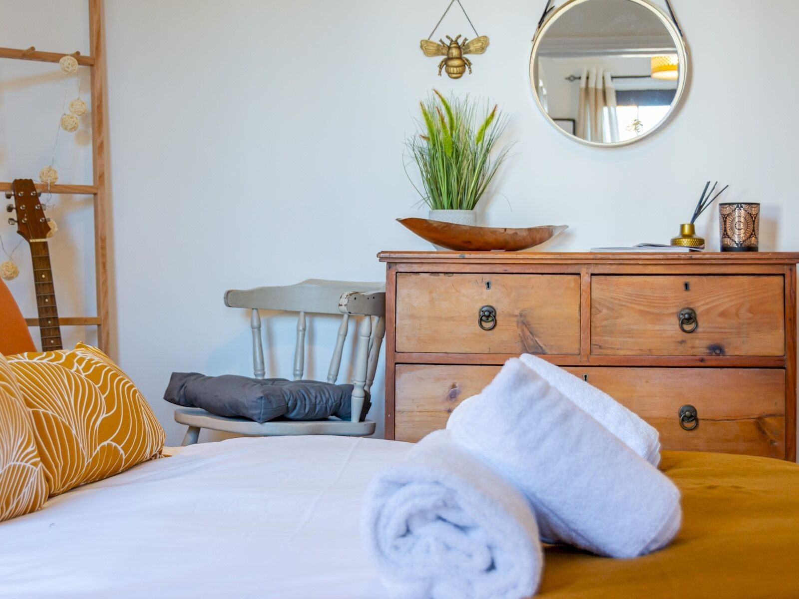 A neatly made bed with a rolled white towel, a wooden dresser with drawers, a small plant on top, a mirror, and a guitar leaning against the wall.