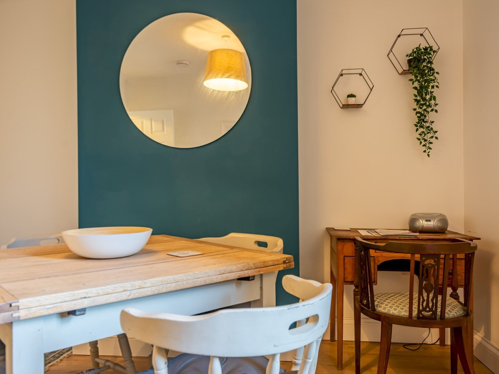 Un coin repas avec une table en bois, des chaises blanches, un miroir rond et un luminaire hexagonal fixé au mur.
