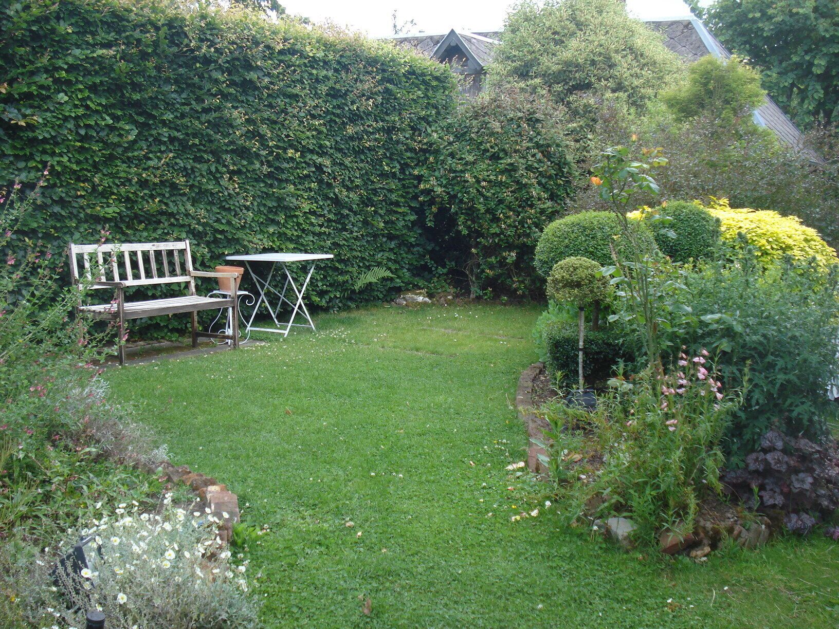 Un jardin avec un banc et une table, entouré de différentes plantes et d’une haie.